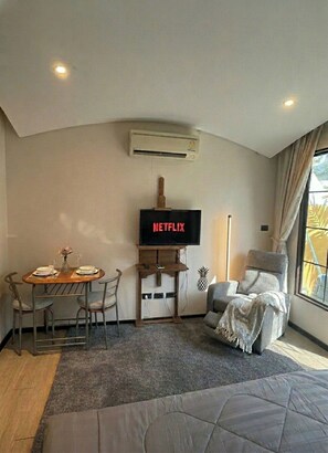Habitación de hotel con una mesa de comedor, sillas, un televisor en un soporte de madera, un sillón reclinable, un ventanal con cortinas y un equipo de aire acondicionado en la pared.