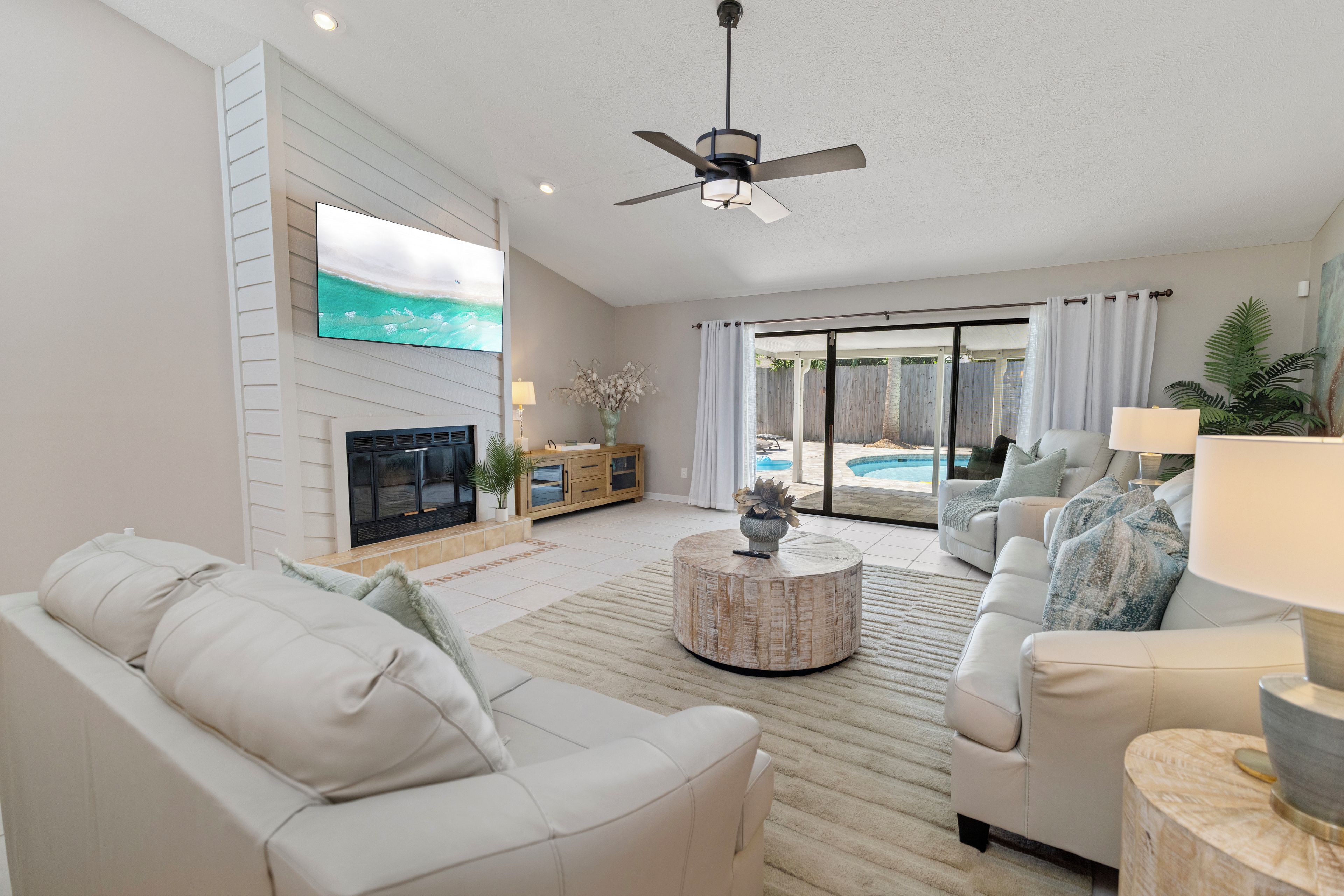 A spacious living room with a flat-screen TV, a fireplace, a ceiling fan, and a view of a pool through glass doors.