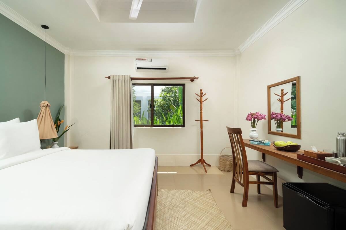 Habitación de hotel con cama, escritorio y silla, ventana con cortinas, perchero, espejo y pequeña mesa con un jarrón de flores.