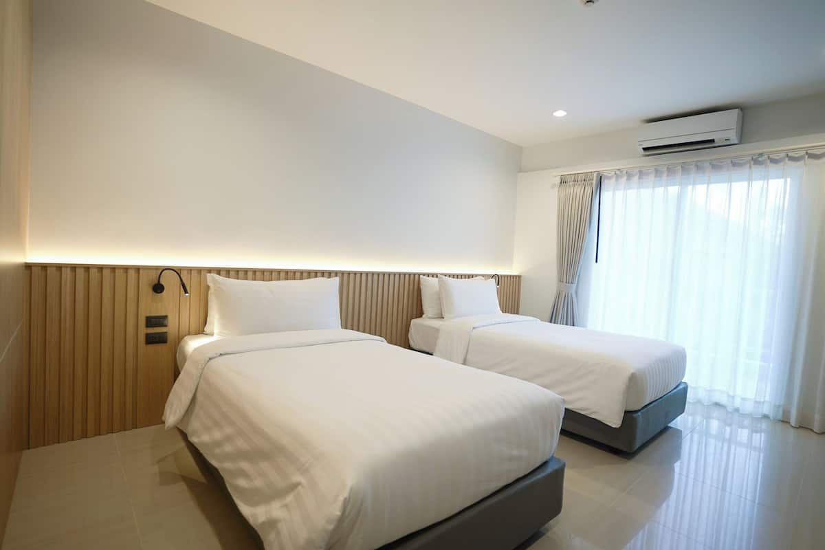 Habitación de hotel con dos camas, un panel de pared de madera, un equipo de aire acondicionado y un ventanal con cortinas traslúcidas.