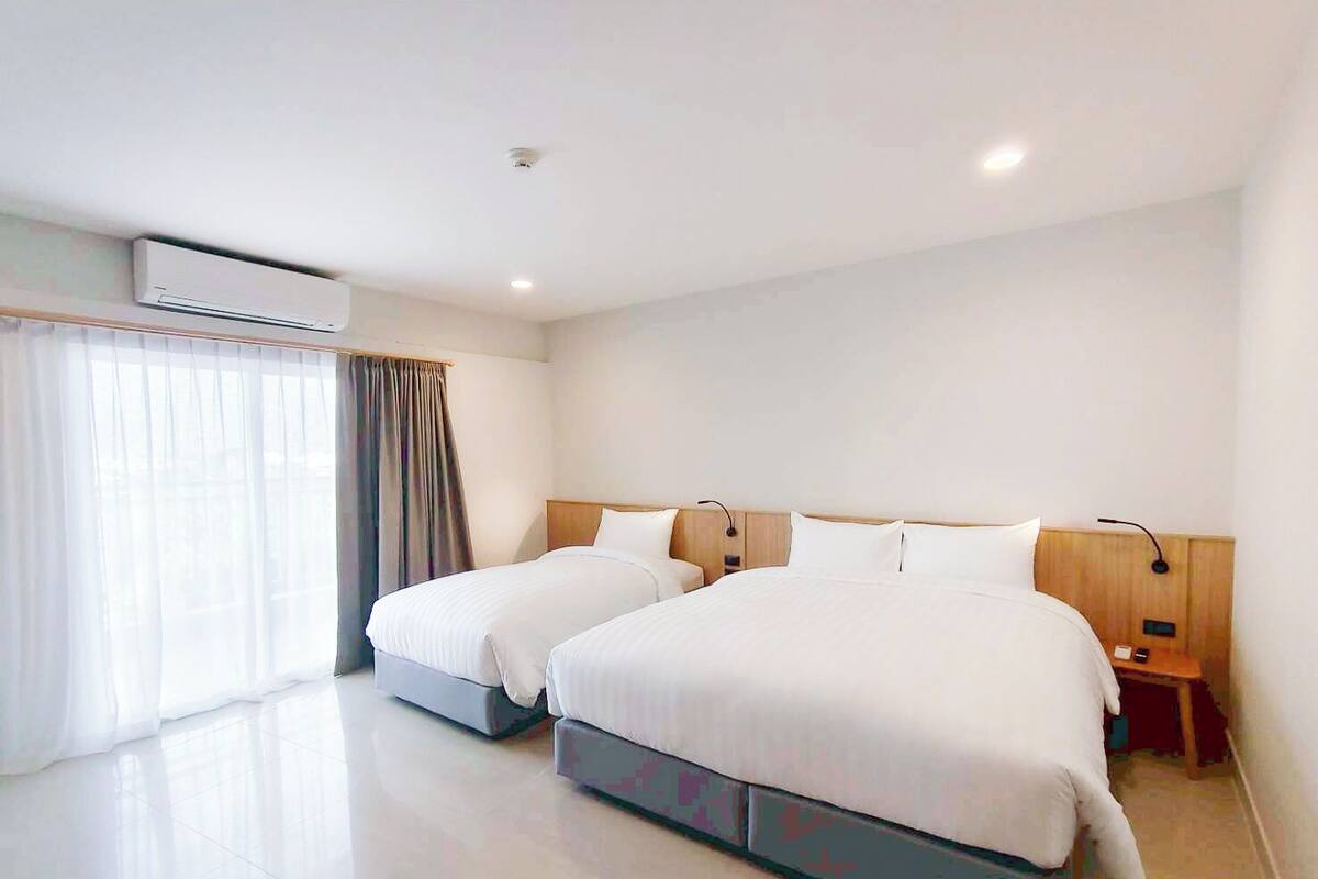 Habitación de hotel con dos camas, un ventanal con cortinas, un equipo de aire acondicionado y una mesita de noche con lámpara.