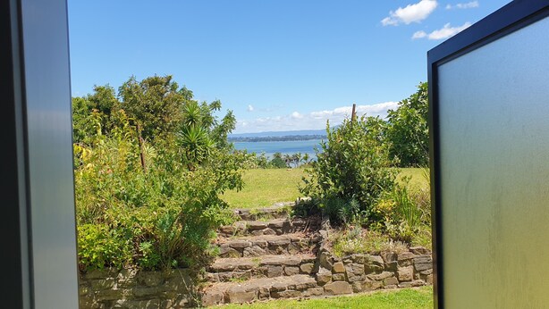 A view through a window of a stone pathway leading to a grassy area with trees and a body of water in the background. - Lazy Daze at Athenree with local hot pools, and only 5 minutes from Waihi Beach (Athenree)