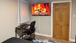 Ett sovrum med en TV som visar en bild av en öppen spis, ett skrivbord, en stol och en dörr av trä.