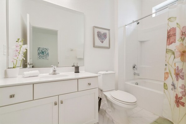 Un baño moderno con lavamanos, inodoro y bañera blancos. Se observa una obra de arte con forma de corazón en la pared y una cortina de ducha con estampado floral.