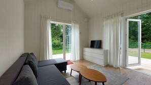 Ett modernt vardagsrum med en soffa, ett soffbord, en tv på ett vitt skåp och stora glasdörrar som leder ut till en trädgård.