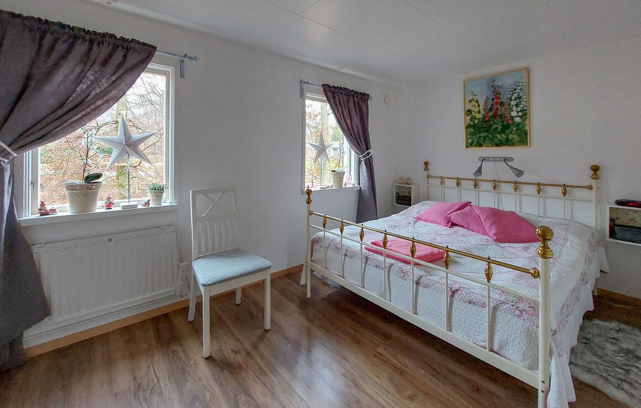 Quarto com cama, cadeira, janela com cortinas, radiador e um quadro na parede.