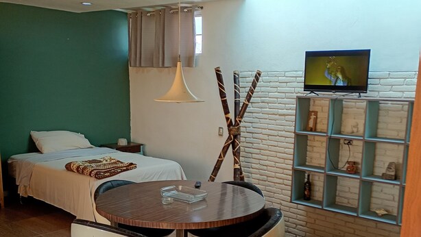 Habitación moderna con una cama, una mesa redonda con dos sillas, un televisor montado en una pared de ladrillo y una lámpara colgante.