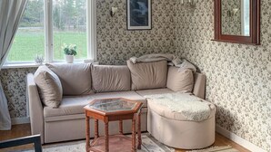 A cozy living room with a beige sectional sofa, a glass-top coffee table, and a round ottoman. A potted plant is on the windowsill, and a framed picture hangs on the wall.
