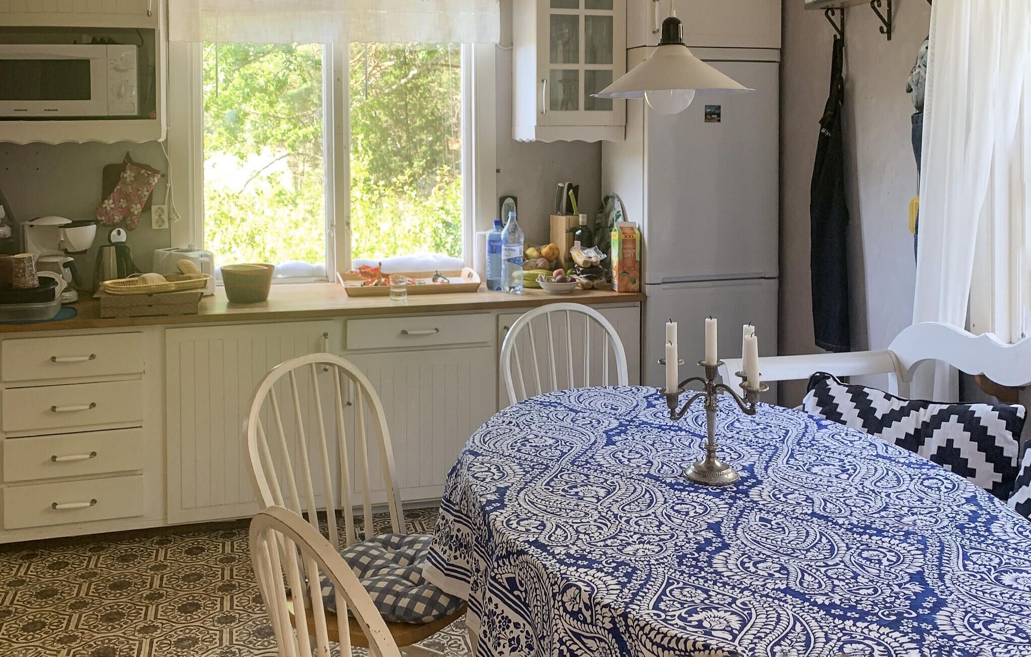 Une cuisine avec des meubles blancs, un four à micro-ondes, un évier et une fenêtre donnant sur un paysage verdoyant. On y trouve une table recouverte d’une nappe à motifs et agrémentée de bougies.