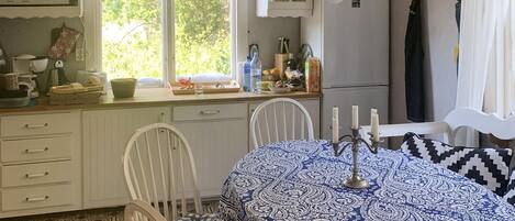 Une cuisine avec des meubles blancs, un four à micro-ondes, un évier et une fenêtre donnant sur un paysage verdoyant. On y trouve une table recouverte d’une nappe à motifs et agrémentée de bougies.