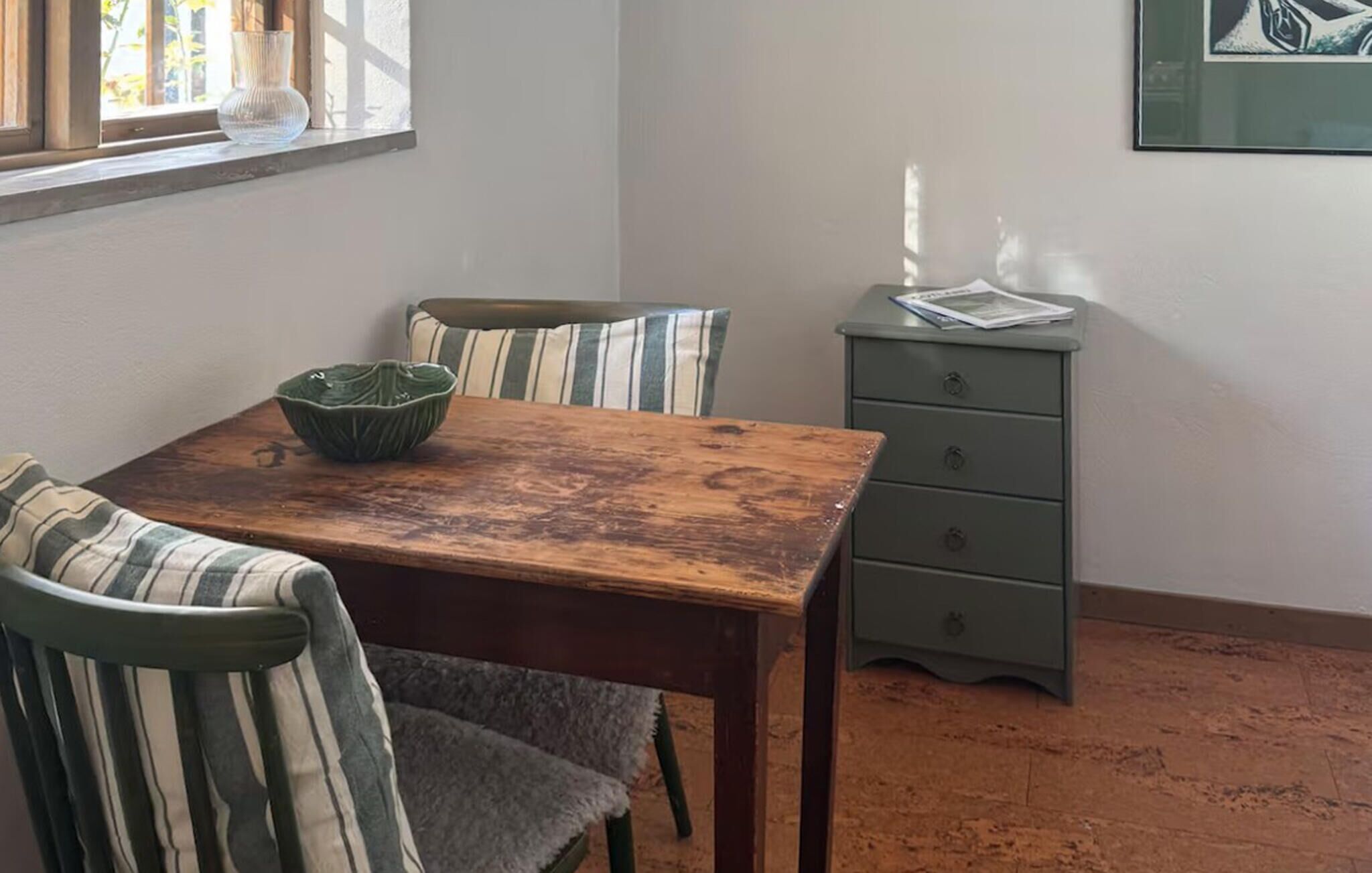 Un comedor con mesa de madera y sillas a rayas, un pequeño aparador verde y un ventanal con un florero.
