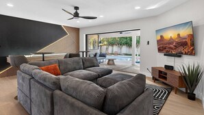 Een moderne woonkamer met een modulaire bank, een plafondventilator, een flatscreen televisie en uitzicht op een zwembad en buitenruimte.