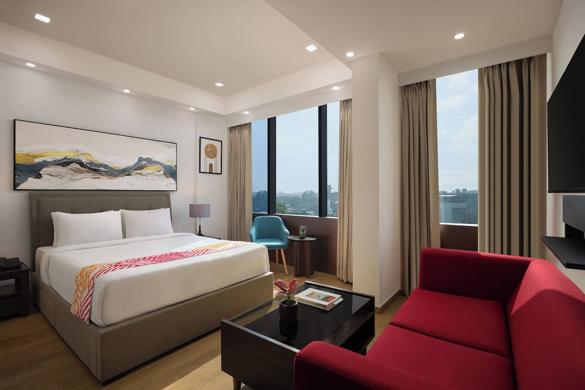 Une chambre d’hôtel moderne avec un grand lit, un canapé rouge, une petite table avec un vase et une vue sur la ville à travers de grandes fenêtres.