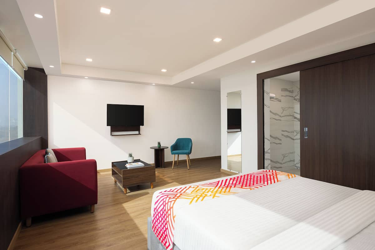 Une chambre d’hôtel moderne avec un grand lit, un canapé rouge, une petite table avec une plante et un téléviseur fixé au mur.