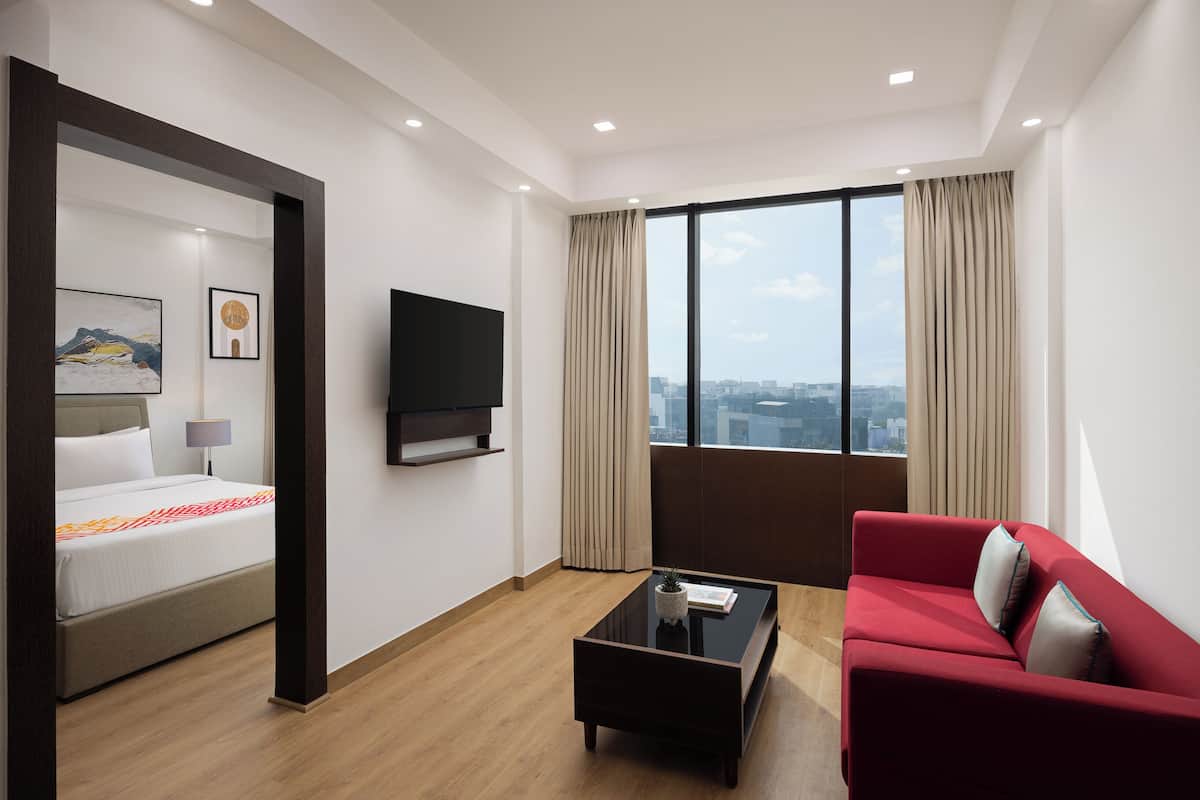 Une chambre d’hôtel moderne, équipée d’un lit, d’un canapé rouge, d’une table basse, d’un téléviseur et d’une grande fenêtre avec des rideaux.