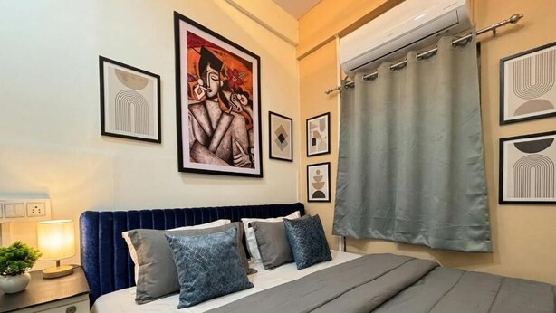 Een slaapkamer met een bed, nachtkastje met lamp, een potplant en lijstjes met kunst aan de muur.