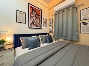 A bedroom with a bed, a nightstand with a lamp, a potted plant, and framed artwork on the wall. - Hotel Elevate Suites (Nagpur)