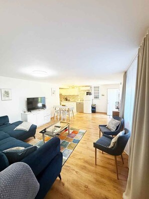 A modern living room with a sofa, armchairs, and a coffee table. There is a television on a cabinet, and a kitchen area is visible in the background. - Holiday flat with terrace (Malinska)