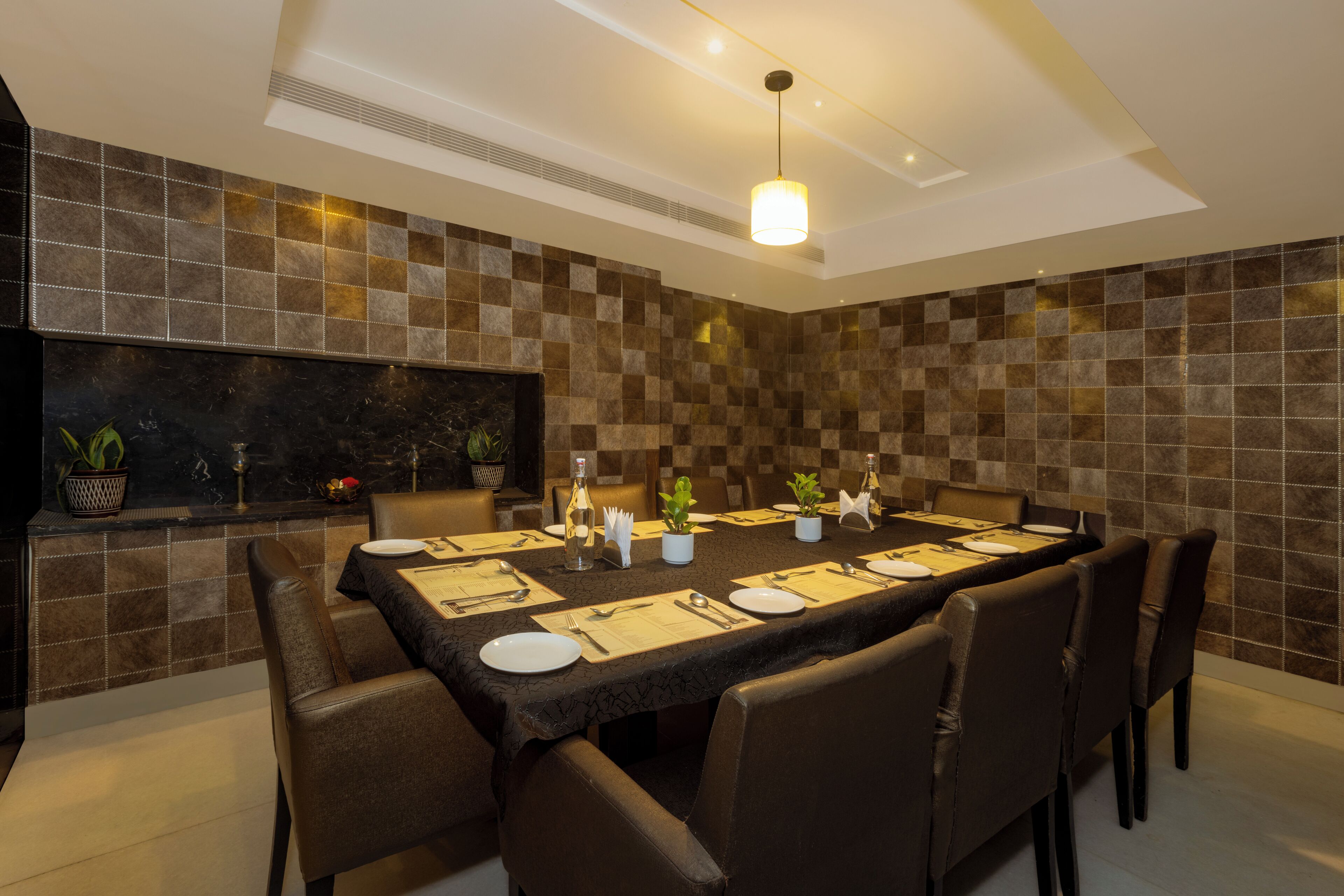 A dining area with a long table set for six, surrounded by chairs, with a tiled wall behind and a ceiling light above.