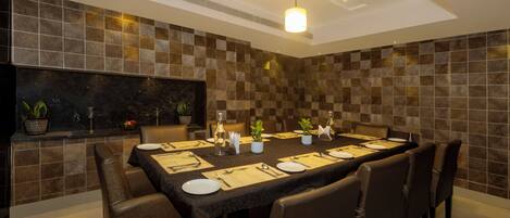 A dining area with a long table set for six, surrounded by chairs, with a tiled wall behind and a ceiling light above.
