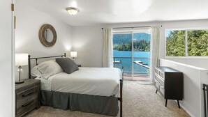 A bedroom with a bed, bedside table, lamp, dresser, and a view of a lake through a large window. - Stylish Escape on Stunning Lake Cavanaugh (Mount Vernon)