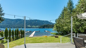 A deck with a view of a lake and dock, surrounded by trees and a grassy area. - Stylish Escape on Stunning Lake Cavanaugh (Mount Vernon)