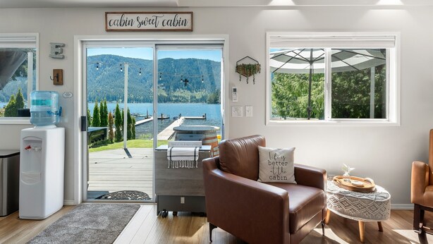 A cabin interior with a large window overlooking a lake and mountains, a water dispenser, a cozy leather sofa, and a small round ottoman. - Stylish Escape on Stunning Lake Cavanaugh (Mount Vernon)