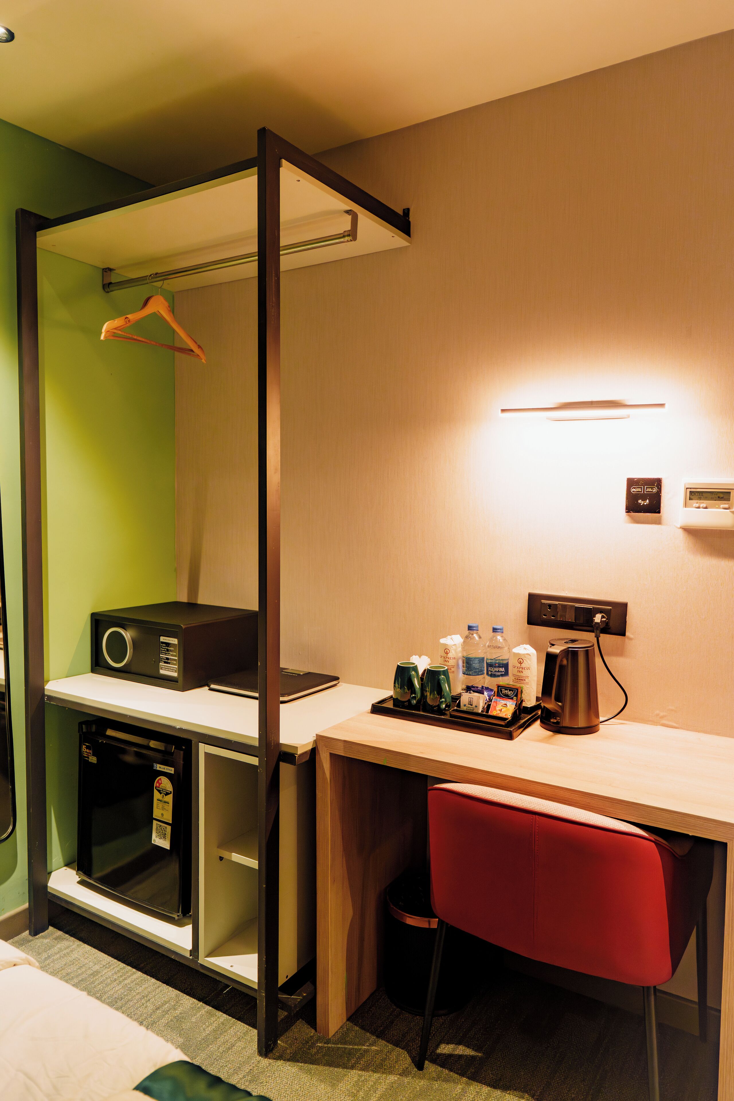 A compact hotel room with a desk, a red chair, a small kitchenette, and a hanging coat rack.