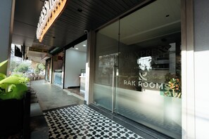 Ingresso di un hotel con tappetino a scacchi, porta in vetro con insegna 'RAK ROOMS' e cartello 'Super Express Laundry'.