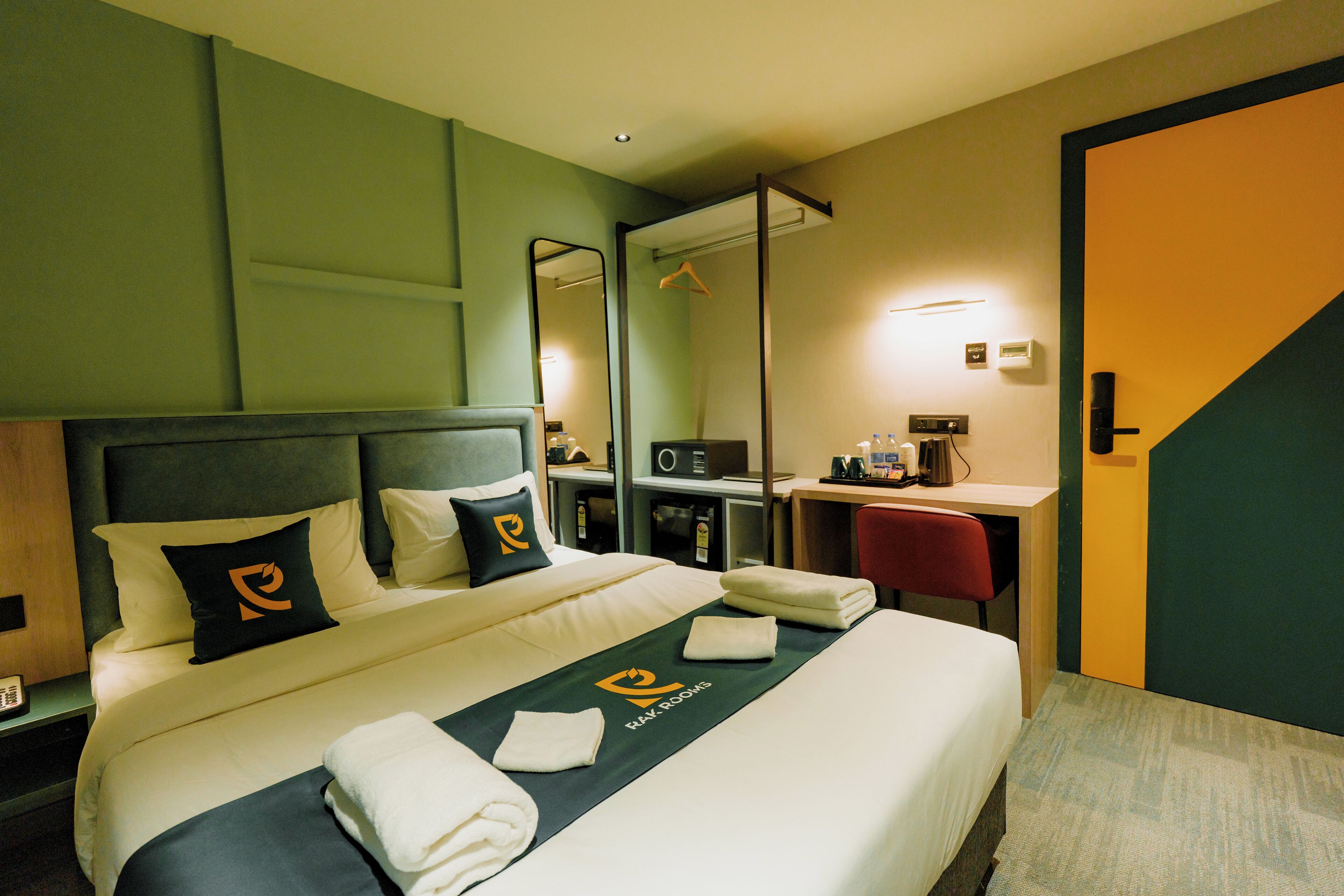 Quarto de hotel com cama, escrivaninha, cadeira, espelho e porta.