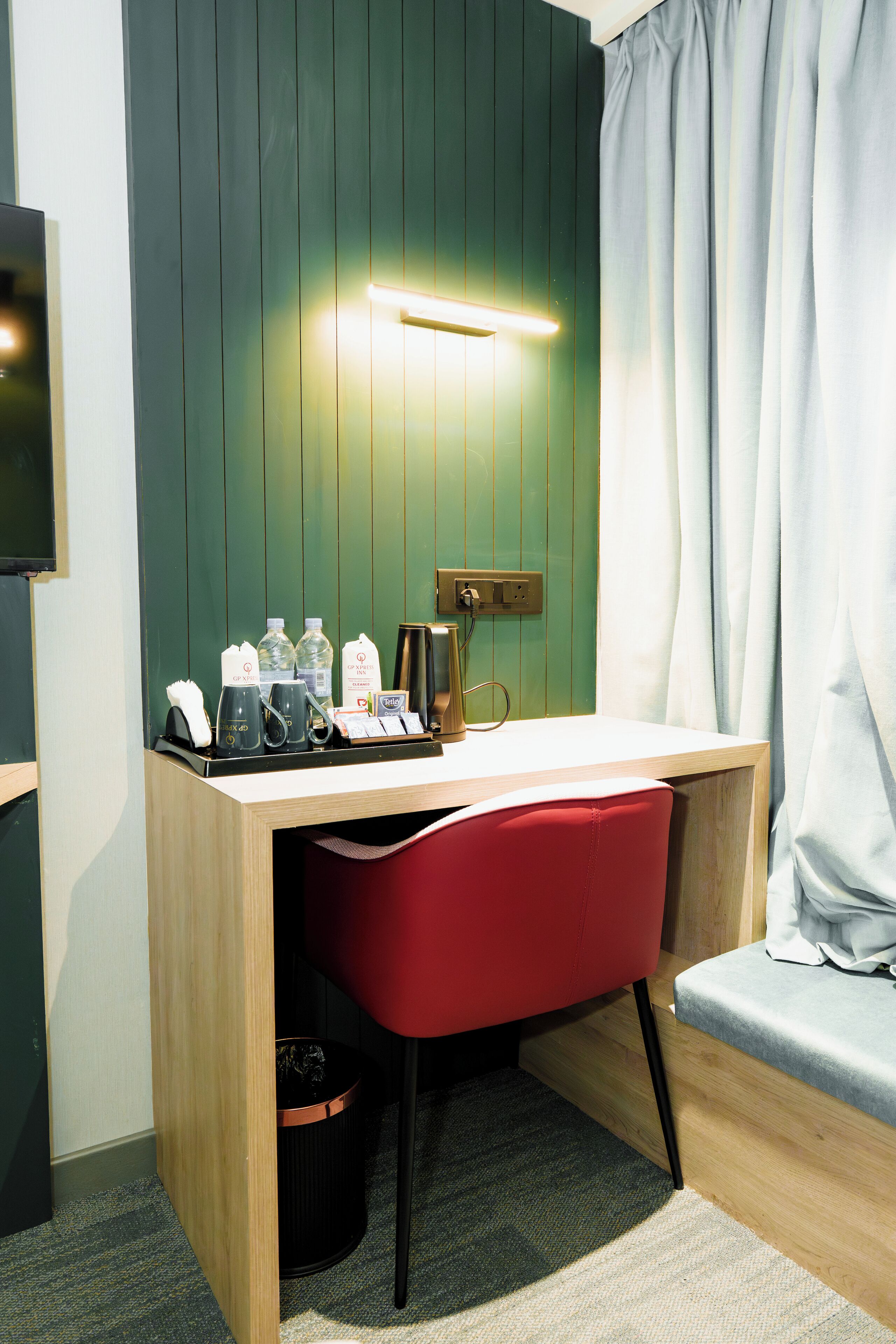 Une chambre d’hôtel compacte, dotée d’un bureau en bois, d’une chaise rouge, d’une petite poubelle, d’une table de chevet et d’un téléviseur fixé au mur.