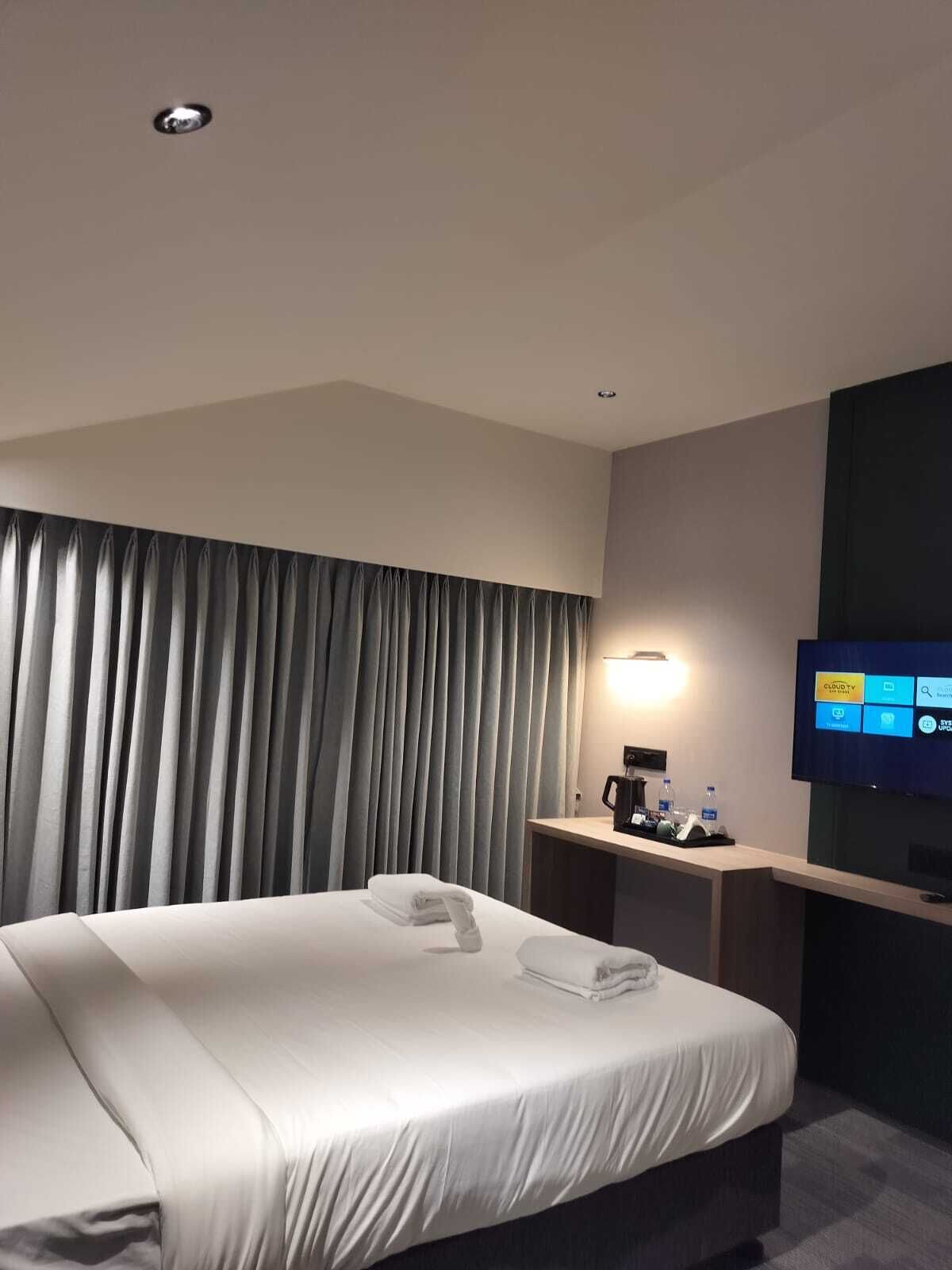 A hotel room with a bed, a desk with a coffee maker, a TV, and a large window with curtains.
