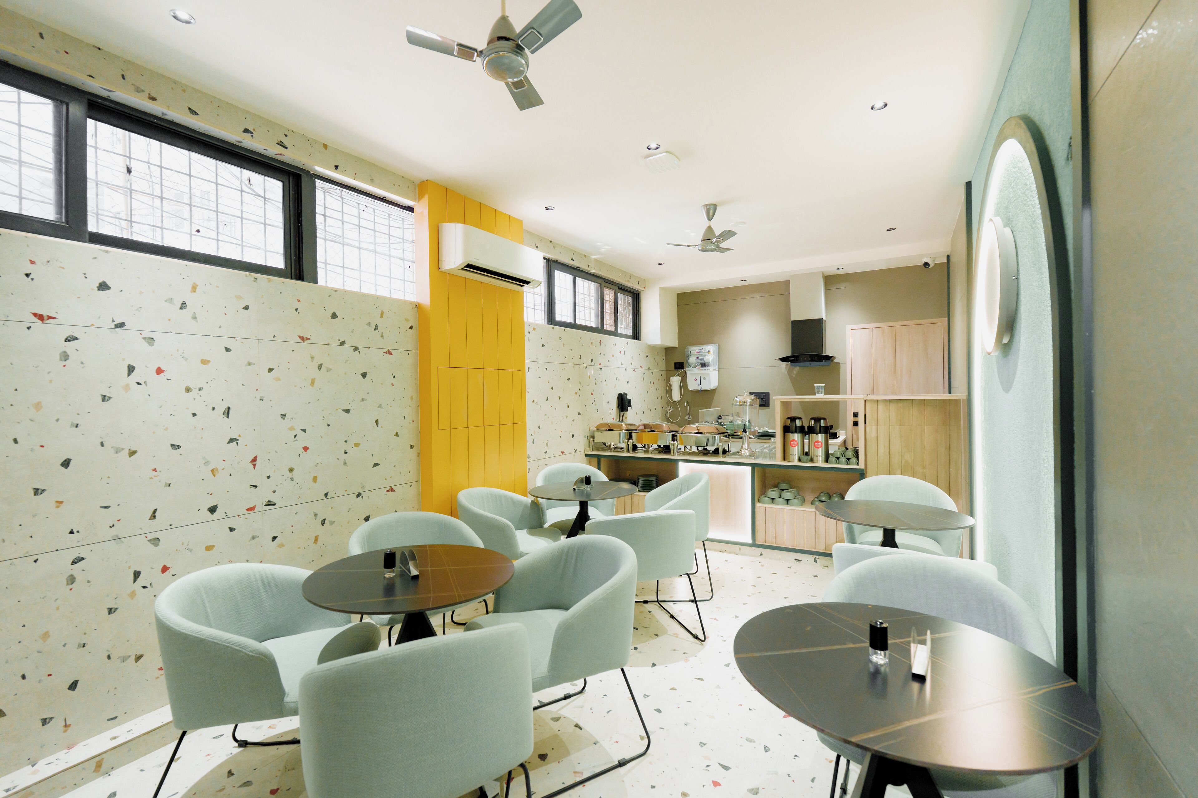 A modern hotel room with a kitchenette, seating area with round tables and chairs, and a large window with a view.