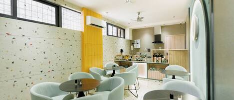 A modern hotel room with a kitchenette, seating area with round tables and chairs, and a large window with a view.