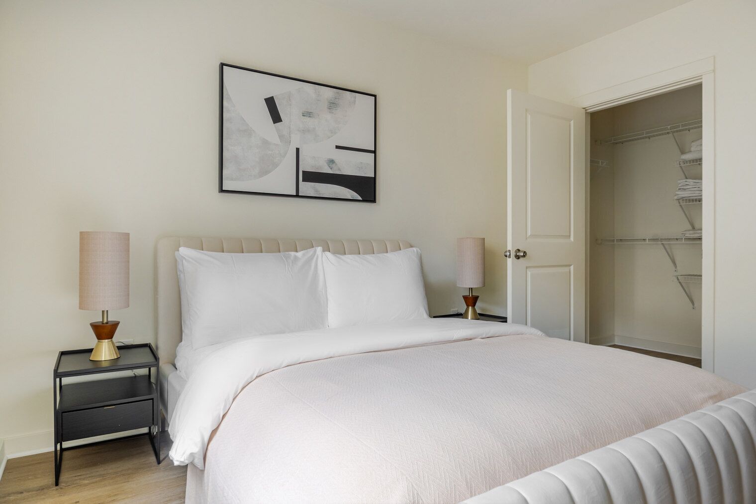 A neatly made bed with white linens, two bedside lamps, a framed abstract artwork above the bed, and a walk-in closet with shelves.
