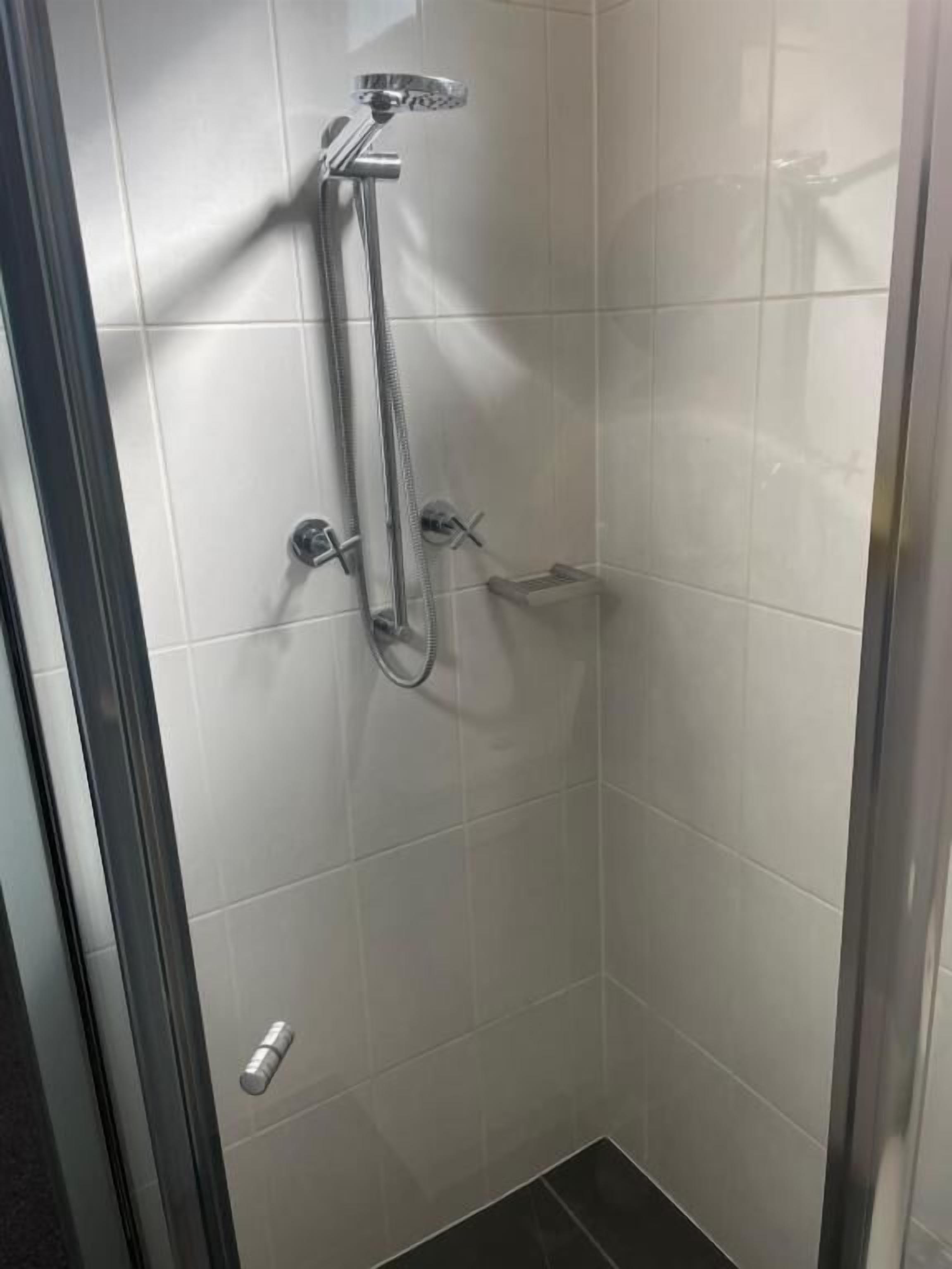 Un petit espace douche fermé, doté d’une porte en verre, d’un pommeau de douche à main et d’un robinet fixé au mur.