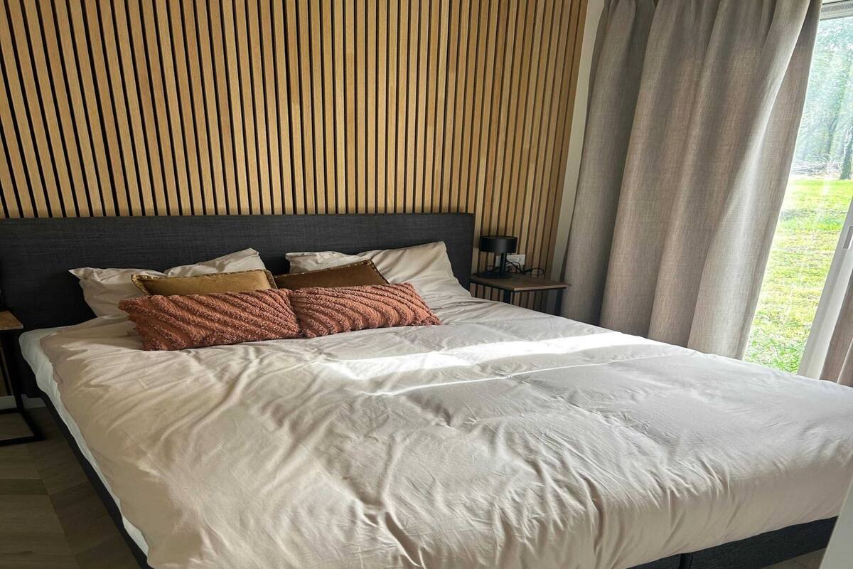 Un lit bien fait, avec une tête de lit grise, une table de chevet avec une lampe, et une vue sur la verdure à l’extérieur.