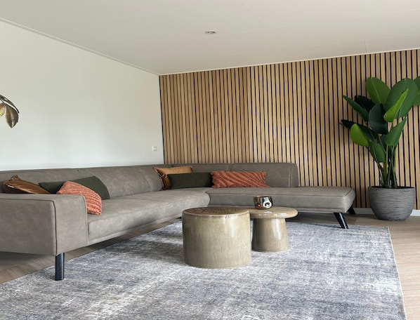 Un salon moderne avec un canapé d’angle gris, deux petites tables rondes, une grande plante en pot et un mur en bois à lattes.