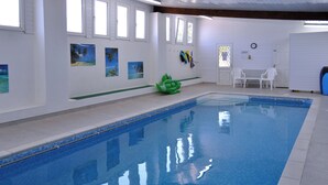 A swimming pool with clear blue water, surrounded by white walls with windows and artwork. - Little Content Farm Cottage (Gunwalloe, near Helston)