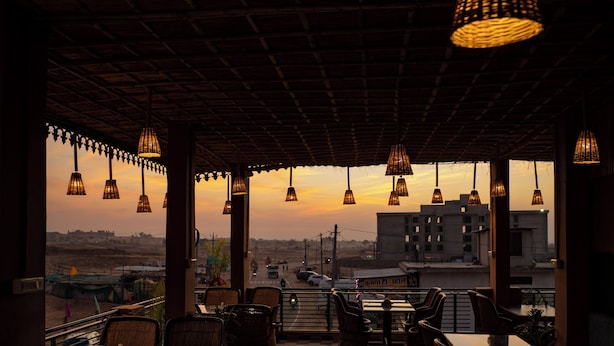 An outdoor seating area with wicker furniture and hanging lights, overlooking a cityscape at sunset. - Pushkar Desert View (Pushkar)
