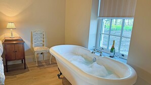 A bathroom with a freestanding bathtub, a wooden nightstand, a chair, and a window with a view of greenery. - Fern Tree Cottage - UK50962 (Kirklington near, Bedale)