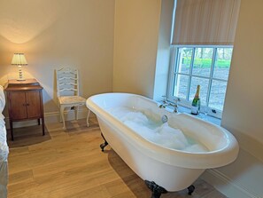 A bathroom with a freestanding bathtub, a wooden nightstand, a chair, and a window with a view of greenery. - Fern Tree Cottage - UK50962 (Kirklington near, Bedale)