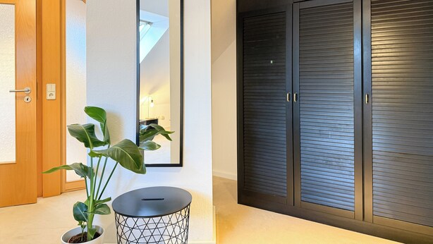 A hallway with a wooden door, a potted plant, a black metal side table, a mirror, and a dark-colored closet with horizontal lines. - Maisonette apartment, large groups, central - NEW (Erfurt)