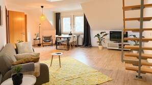 A modern living room with a sofa, a coffee table, a dining area with chairs, a TV, and a staircase. - Maisonette apartment, large groups, central - NEW (Erfurt)