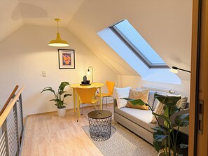 Et hyggeligt værelse med ovenlys, et spisebord med stole, en sofa og et lille rundt bord med lampe.
