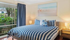 Een slaapkamer met een groot bed, een plafondventilator, een raam met gordijnen, een bedlampje en een schilderij met een strandthema aan de muur.