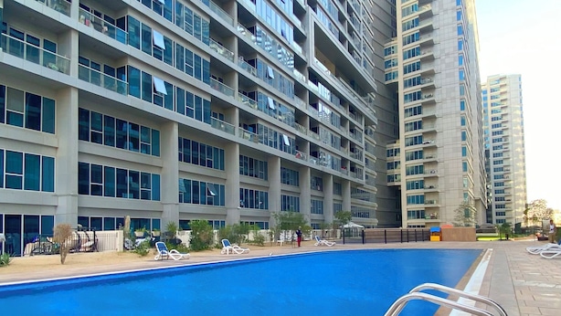 A swimming pool with a clear blue water surface, surrounded by a tiled deck and a metal ladder. In the background, there are high-rise buildings with multiple balconies and large windows. - SkyCourt Tower By Hostiva (Dubai)