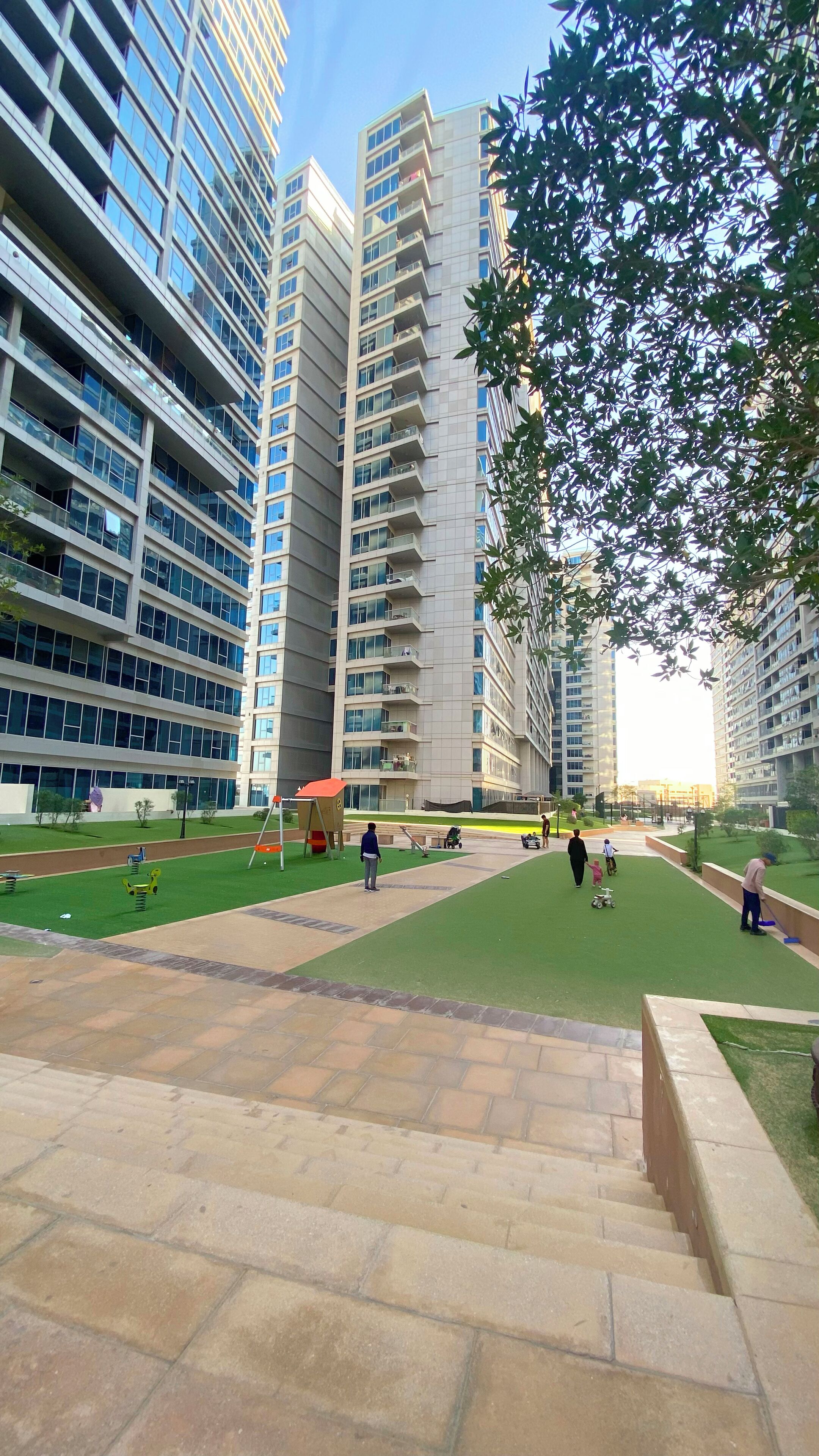 A modern urban park with green lawns, playground equipment, and tall residential buildings in the background.