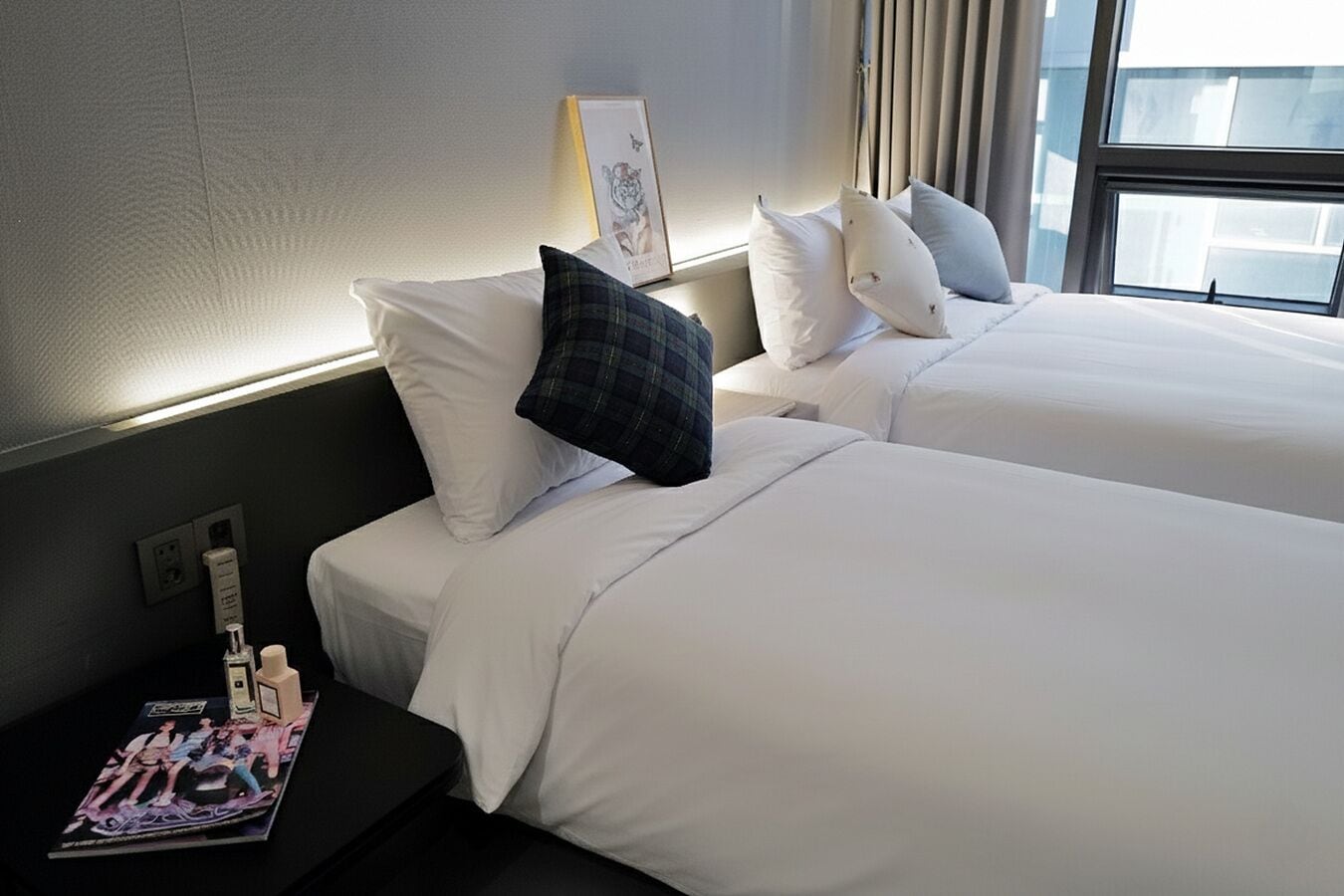 Een hotelkamer met twee bedden, witte beddengoed, een nachtkastje met toiletartikelen, een lijst met een afbeelding en een raam met gordijnen.