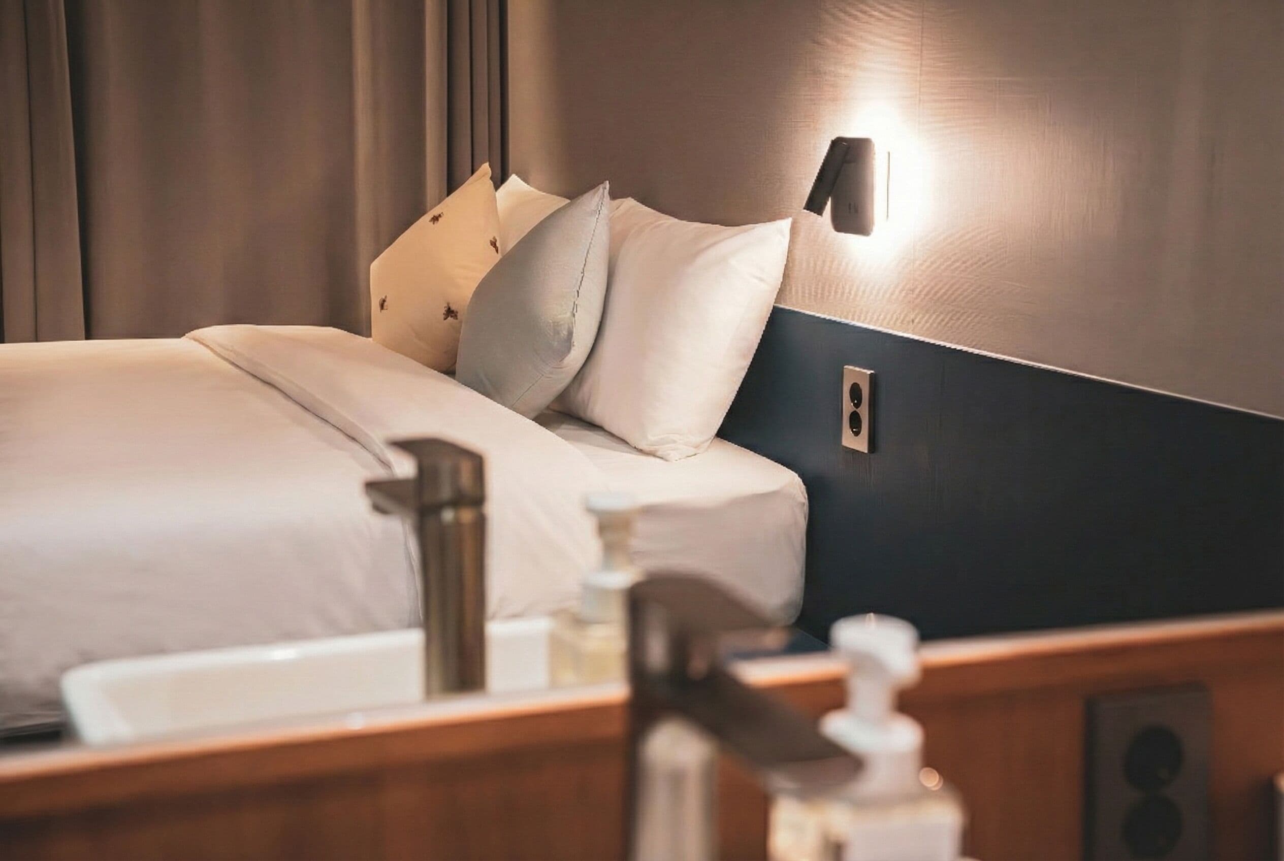 Una cama bien hecha con sábanas y almohadas blancas, una mesita de noche con una botella de jabón de manos y una lámpara de pared.